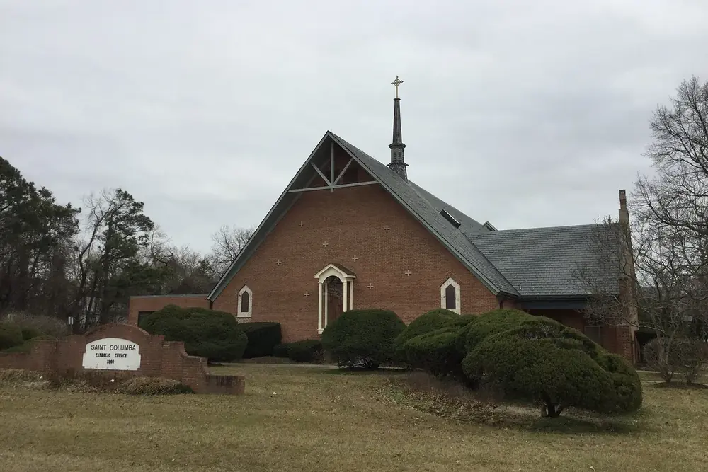 Brick Saint Columba church with a slender spire behind trimmed shrubs in Oxon Hill, Maryland